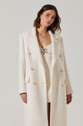 A woman is wearing a long, cream-colored, double-breasted coat with large buttons. The coat features wide lapels and a tailored fit. Underneath, she has a cream-colored top with a lace-up detail at the neckline.